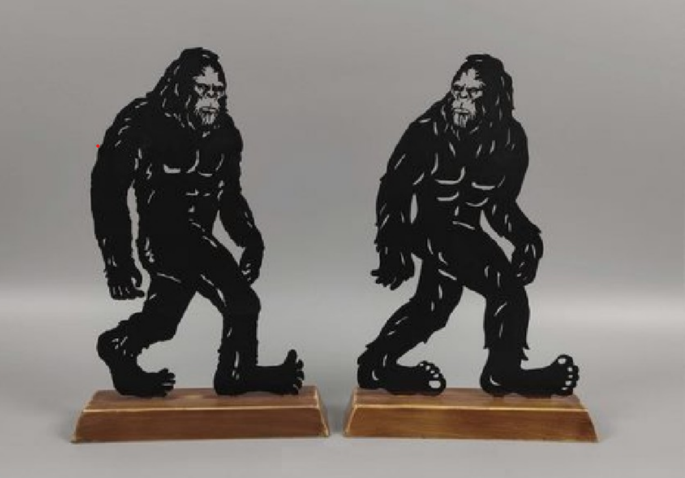 Two black metal cutout sculptures of a Bigfoot figure on wooden bases.