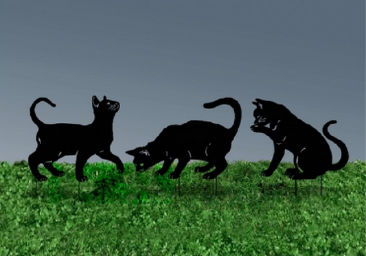 Silhouette yard stakes of cats on a grassy field with a gray background.  One looking up, one sniffing the ground, and one licking its paw.